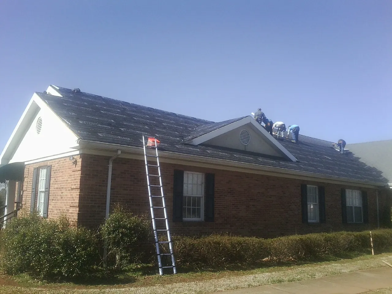 Crew tearing shingles off a brick ranch roof with a ladder set — Metal Roof Repair work by our crew in Mooreland