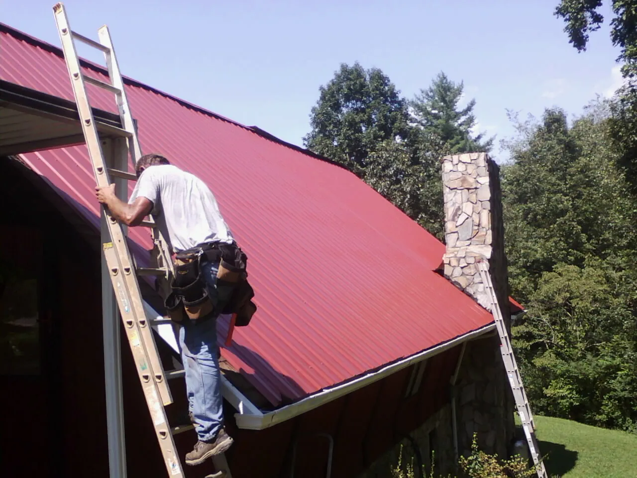 Roofer on a ladder beside a red metal roof and stone chimney — Soffit & Fascia Repair by Roofing Brothers in Mooreland